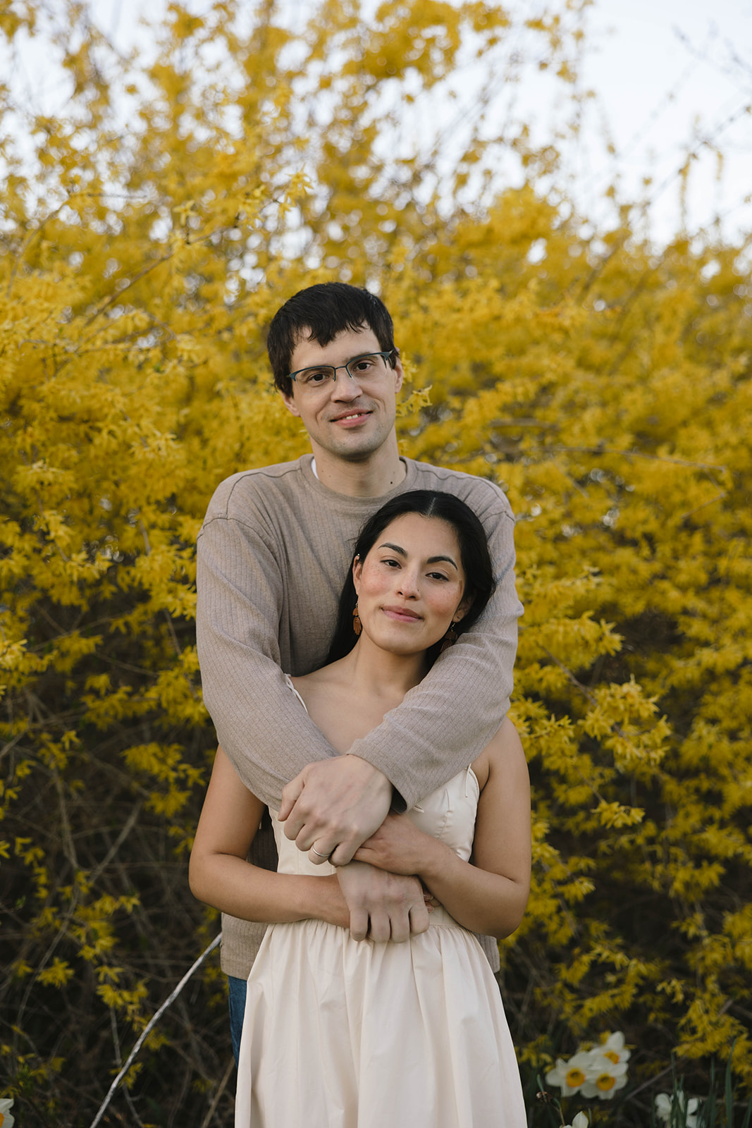 Corey embracing Gaby in front of a yellow blooming bush